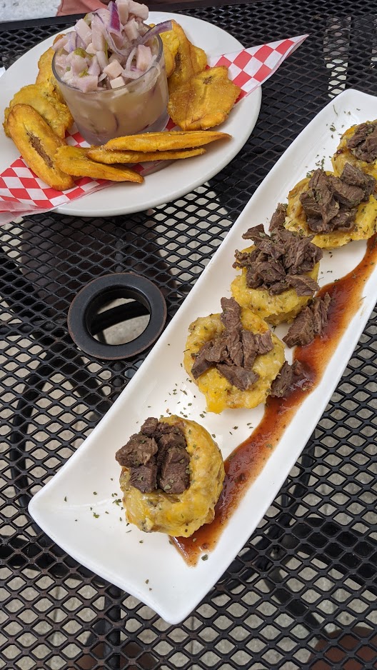Tostones topped with steak alongside a cup of ceviche on a patio table