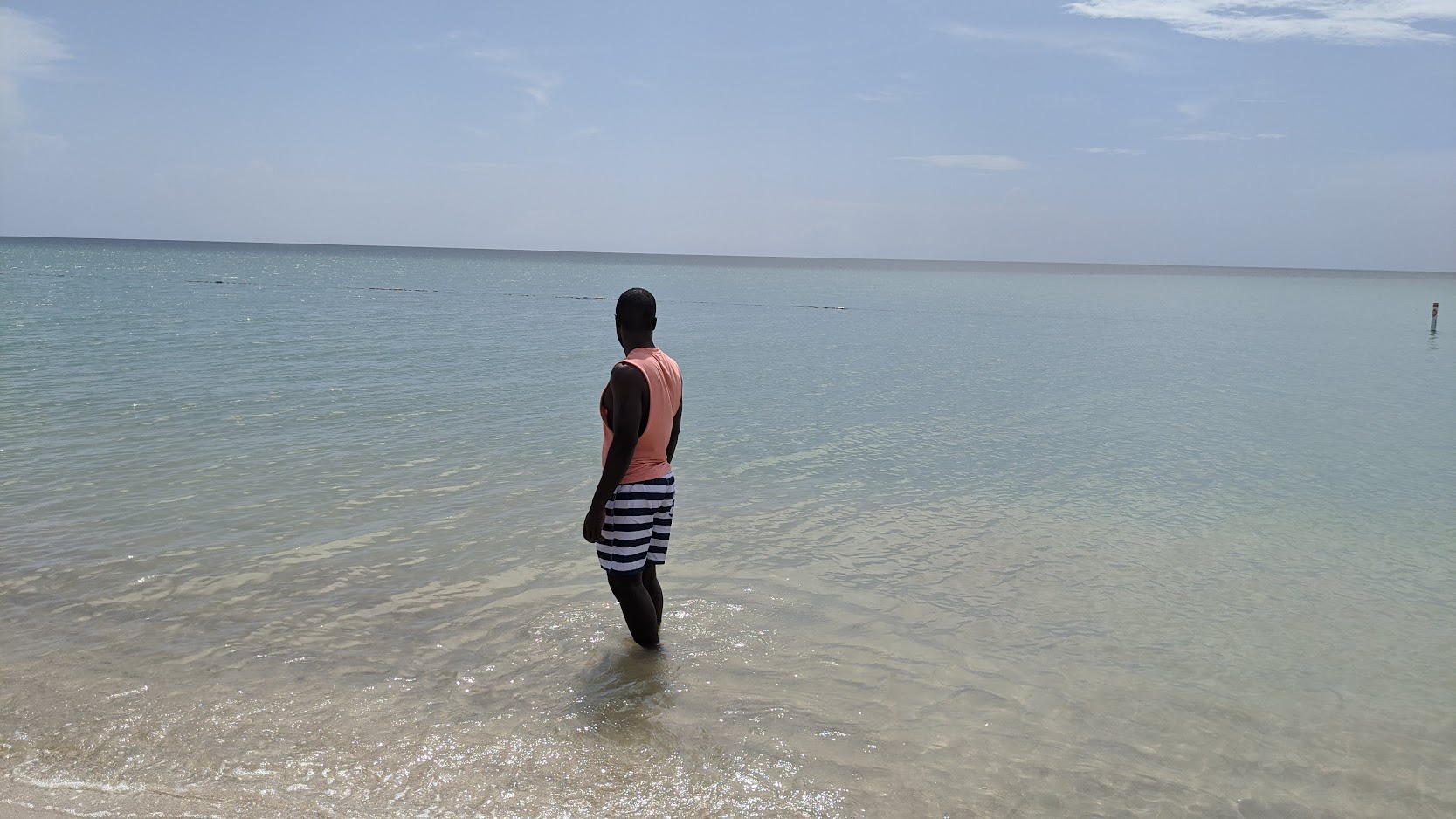 Man in a coral tank top standing ankle-deep in calm, glassy Caribbean water