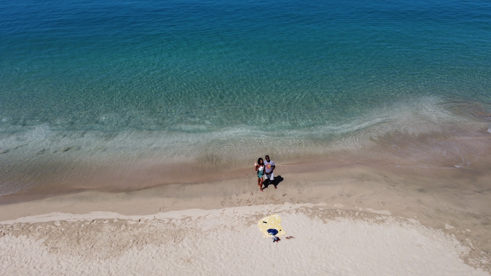 Drone view of a couple standing at the waterline of a wide turquoise beach