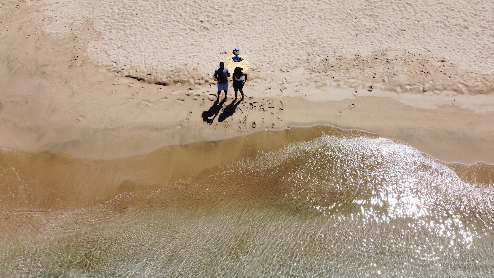 Aerial view of three people standing on the sand as a wave rolls in